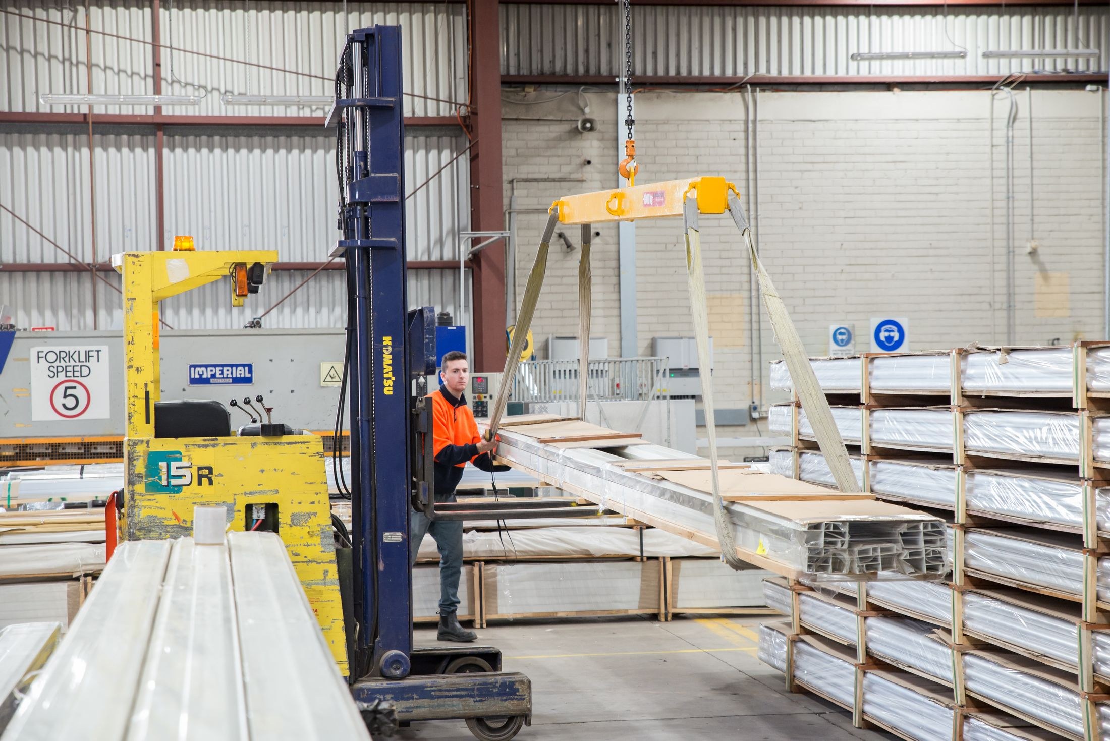 Worker handling wrapped metal profiles with lifting equipment beside an industrial forklift