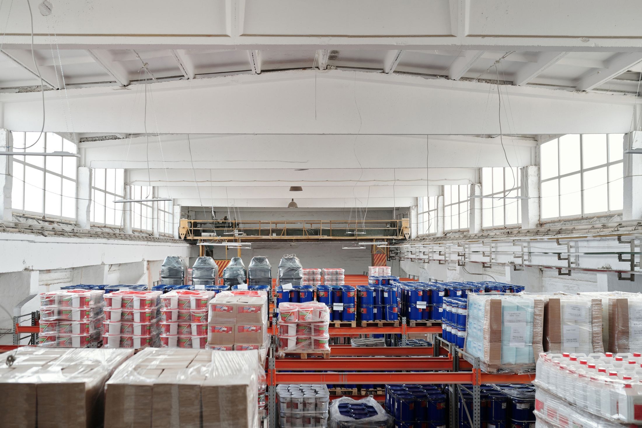 Stocked warehouse racks holding wrapped cartons and pails below a bright industrial roof